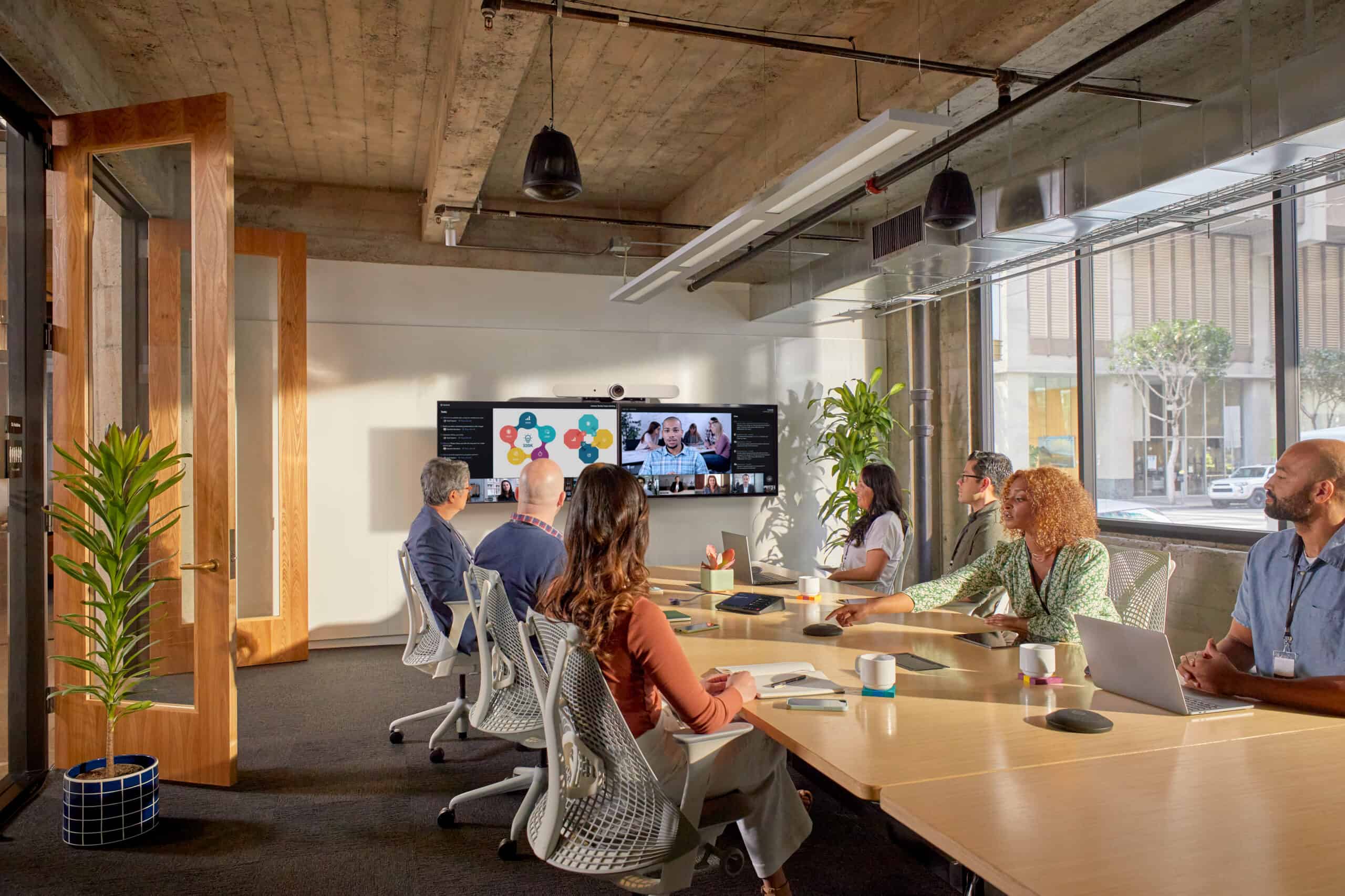 A group in a modern office joins a video meeting, surrounded by large windows, plants, and office supplies.