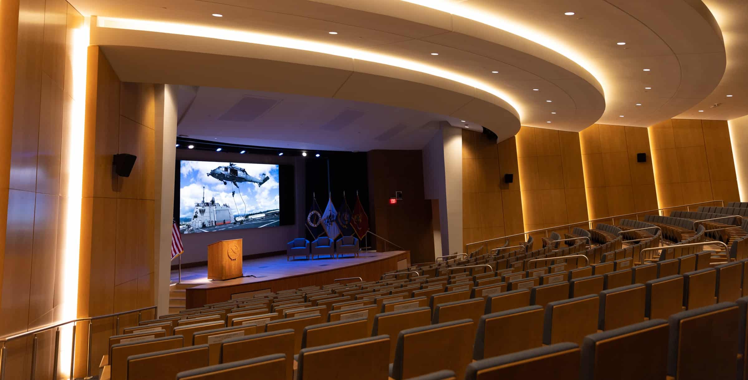 A modern auditorium with curved empty seats, stage with podium, chairs, screen showing helicopter & ship; flags & soft lighting.