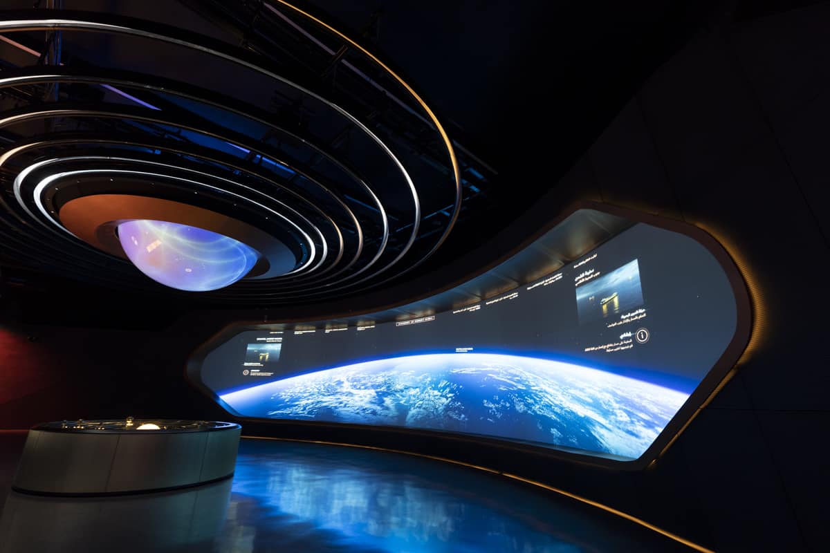 A futuristic exhibit with a curved Earth display, glowing ring lights, and a digital control table in dim lighting.