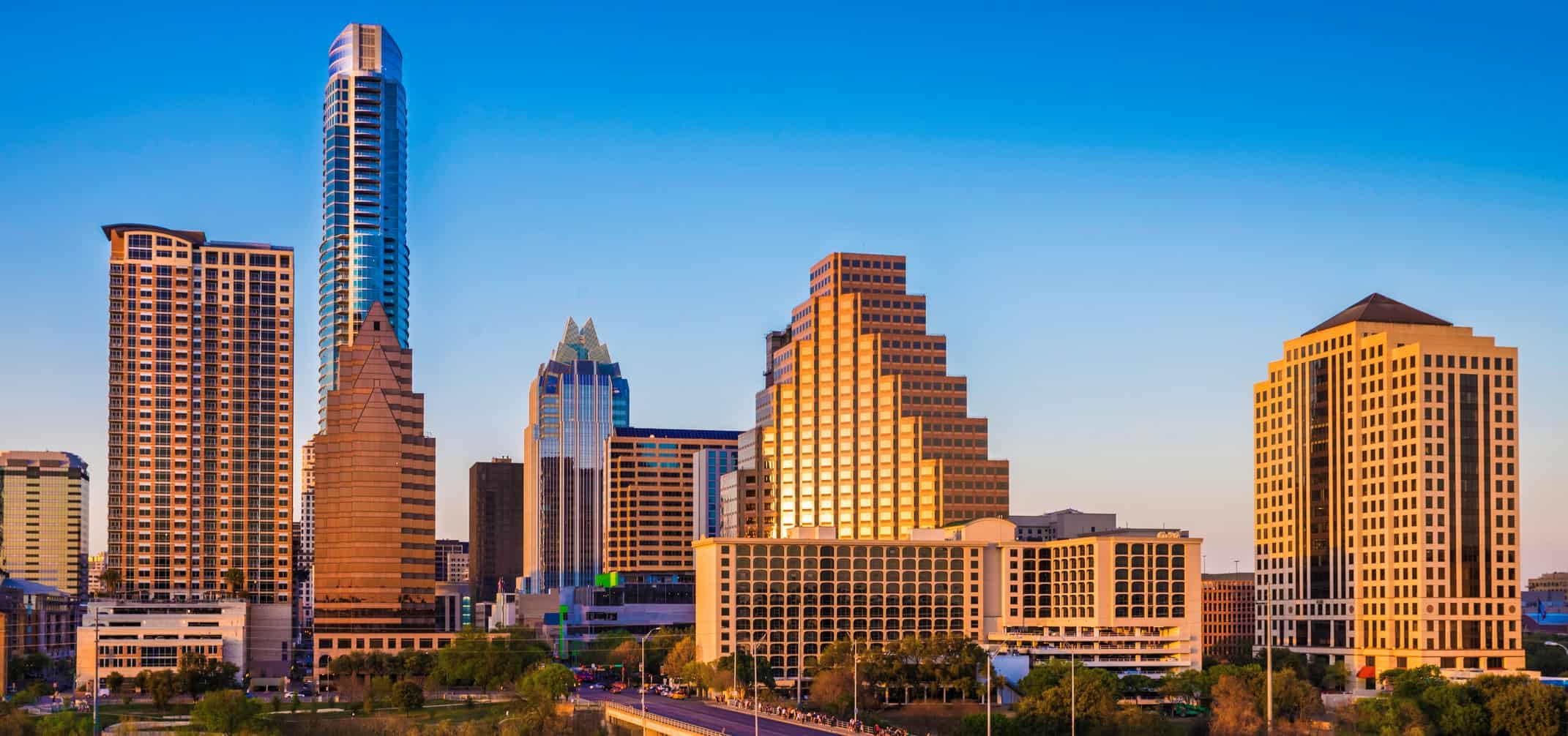 City skyline with diverse skyscrapers under clear blue sky; sunlit reflection. Road and greenery in foreground.