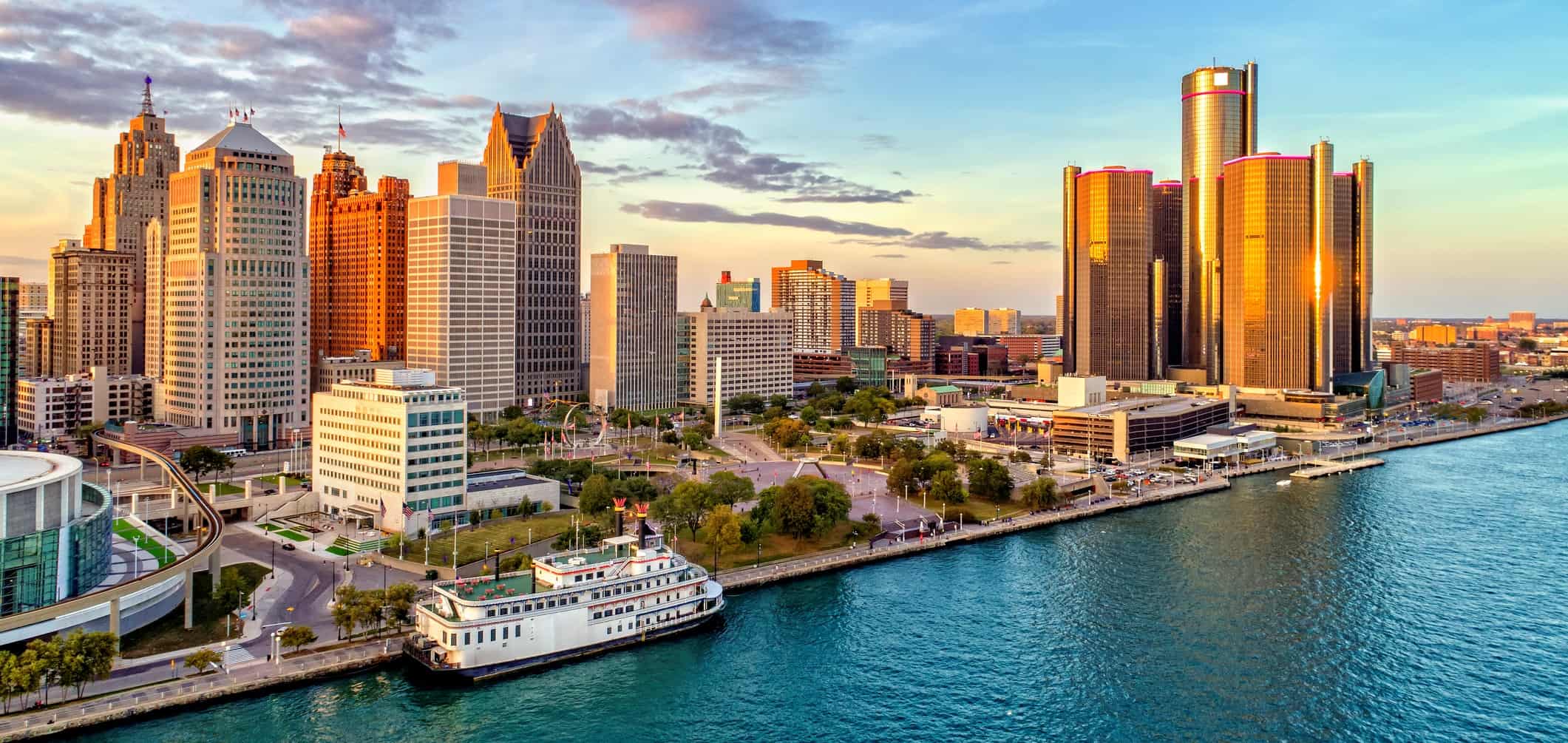 Aerial view: city skyline at sunset, skyscrapers & historic buildings; river with docked ferry, sky blends clouds & golden light.