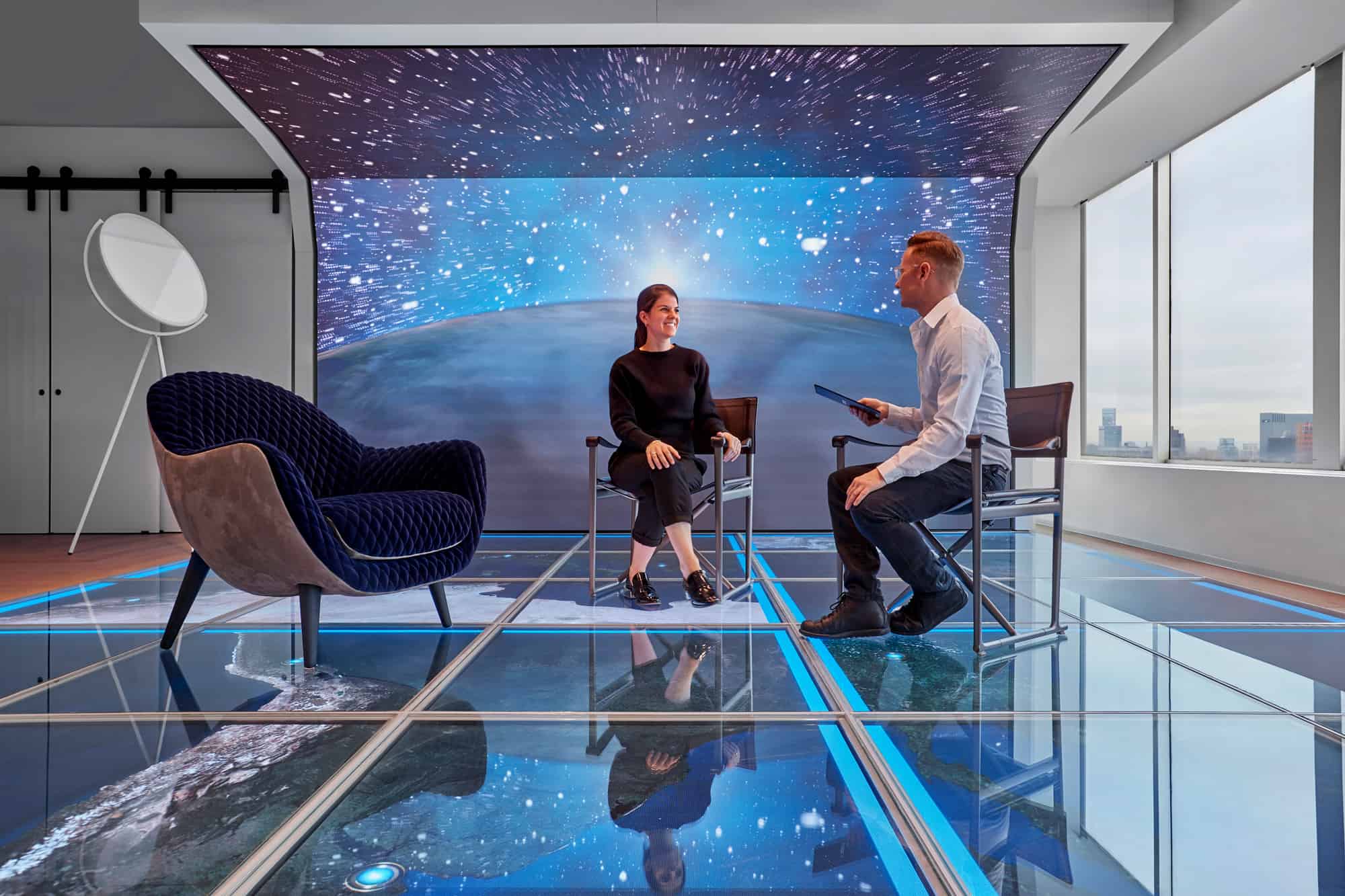Two people sit in a modern room with a space-themed backdrop, glass floor, and city views. One holds a tablet near a large spherical lamp.