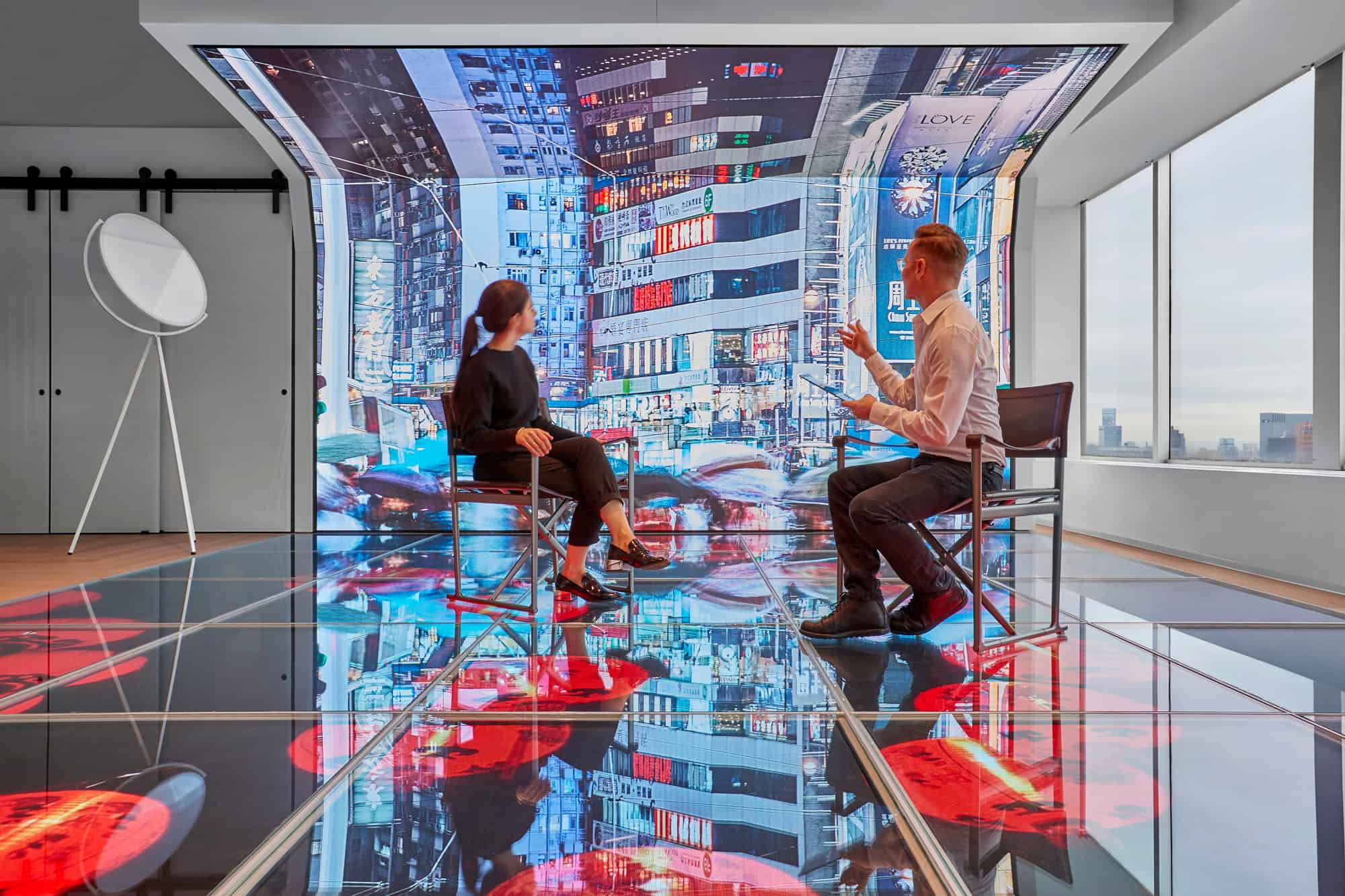 Two people sit on a reflective floor facing each other, digital screen shows city lights; modern room with cityscape window.