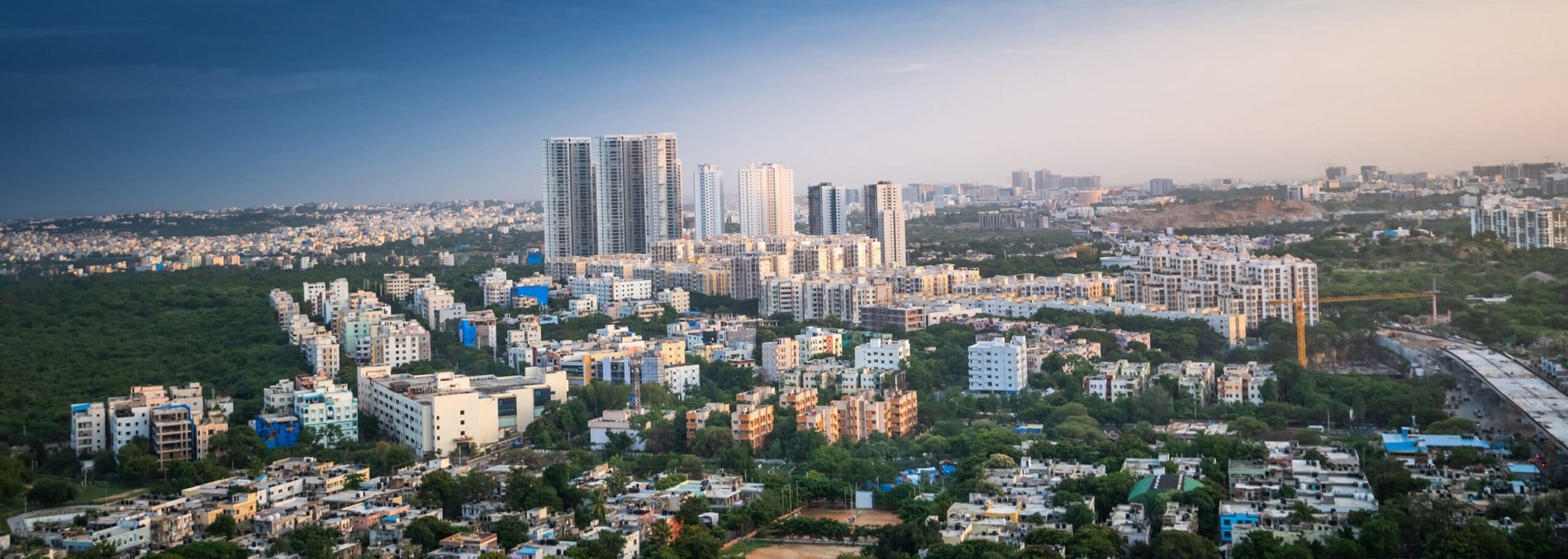 Aerial view of a cityscape with skyscrapers, greenery, and a clear blue sky. The skyline extends beautifully into the distance.