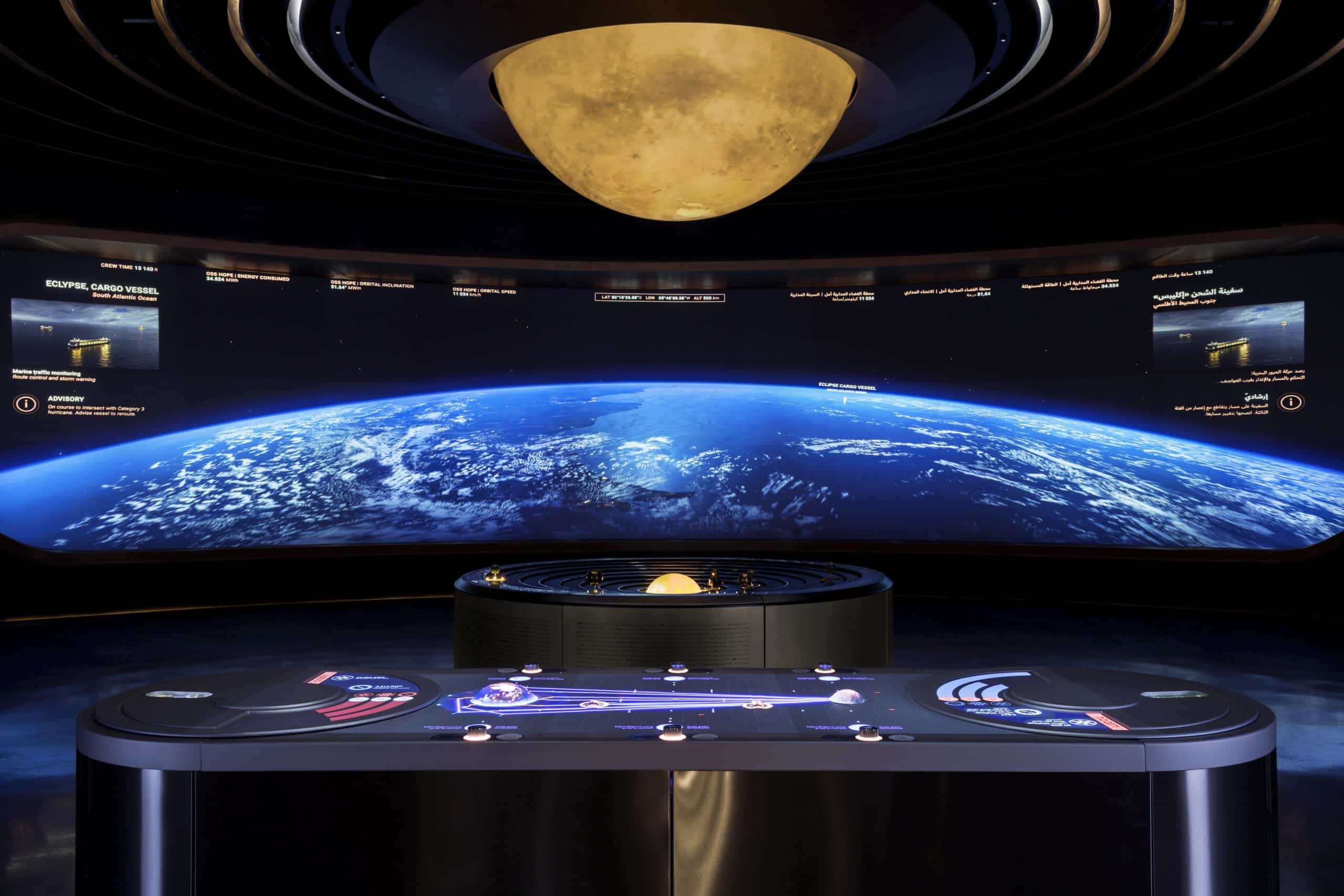 Futuristic room: large curved screen shows Earth; central control panel with solar system model; glowing circular ceiling light.