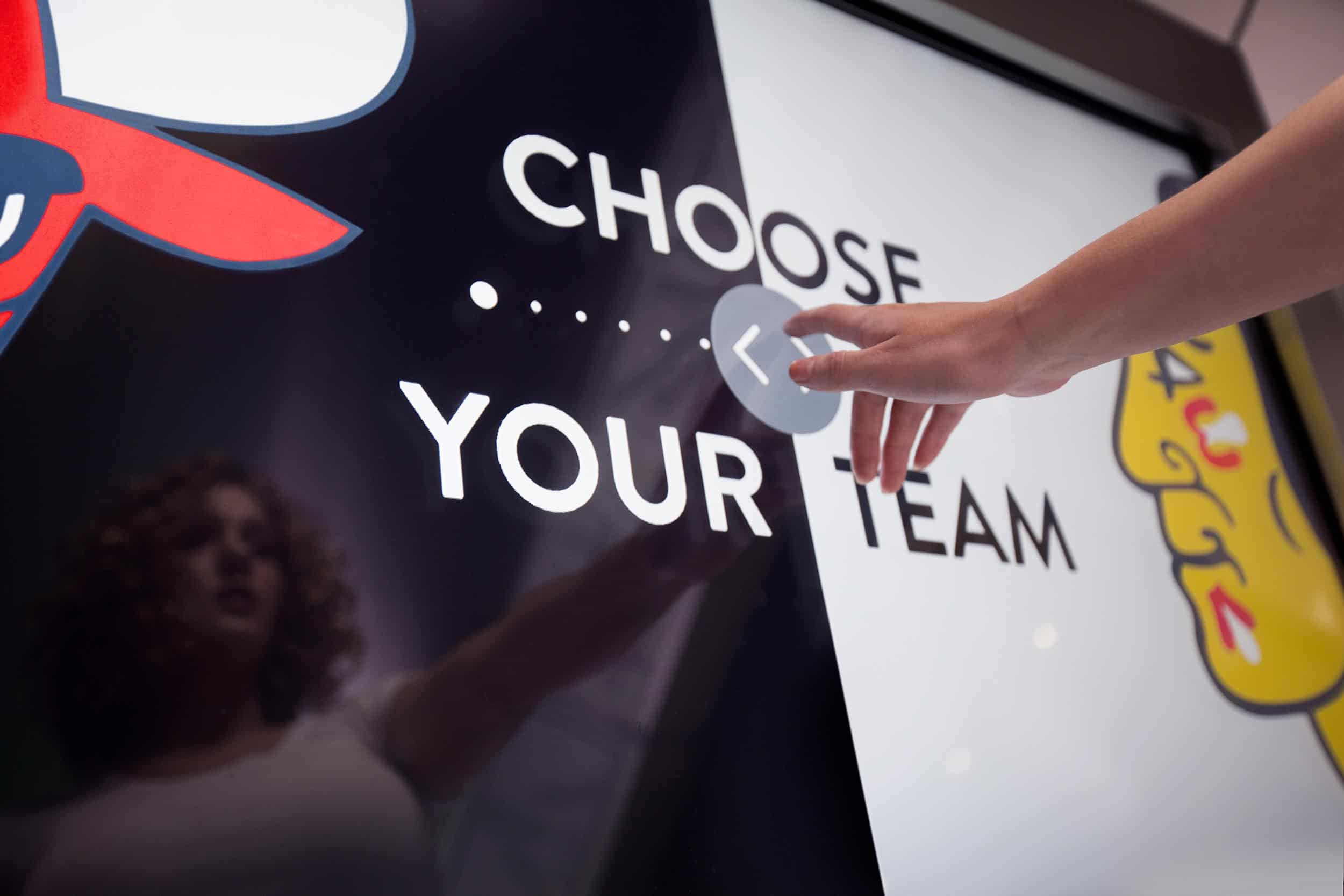 Person at touchscreen: Choose Your Team. Screen shows red wing, yellow element; reflection visible on the left.