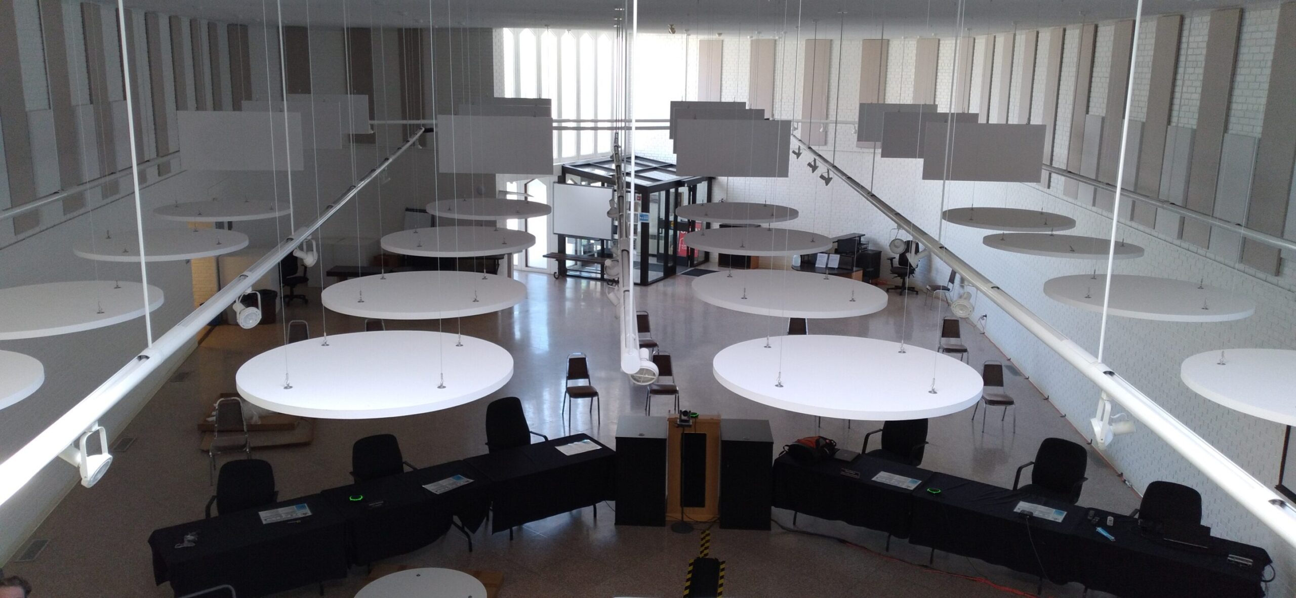 Aerial view: modern, empty conference room; circular lights, black tables & chairs, glass wall, natural light from large windows.