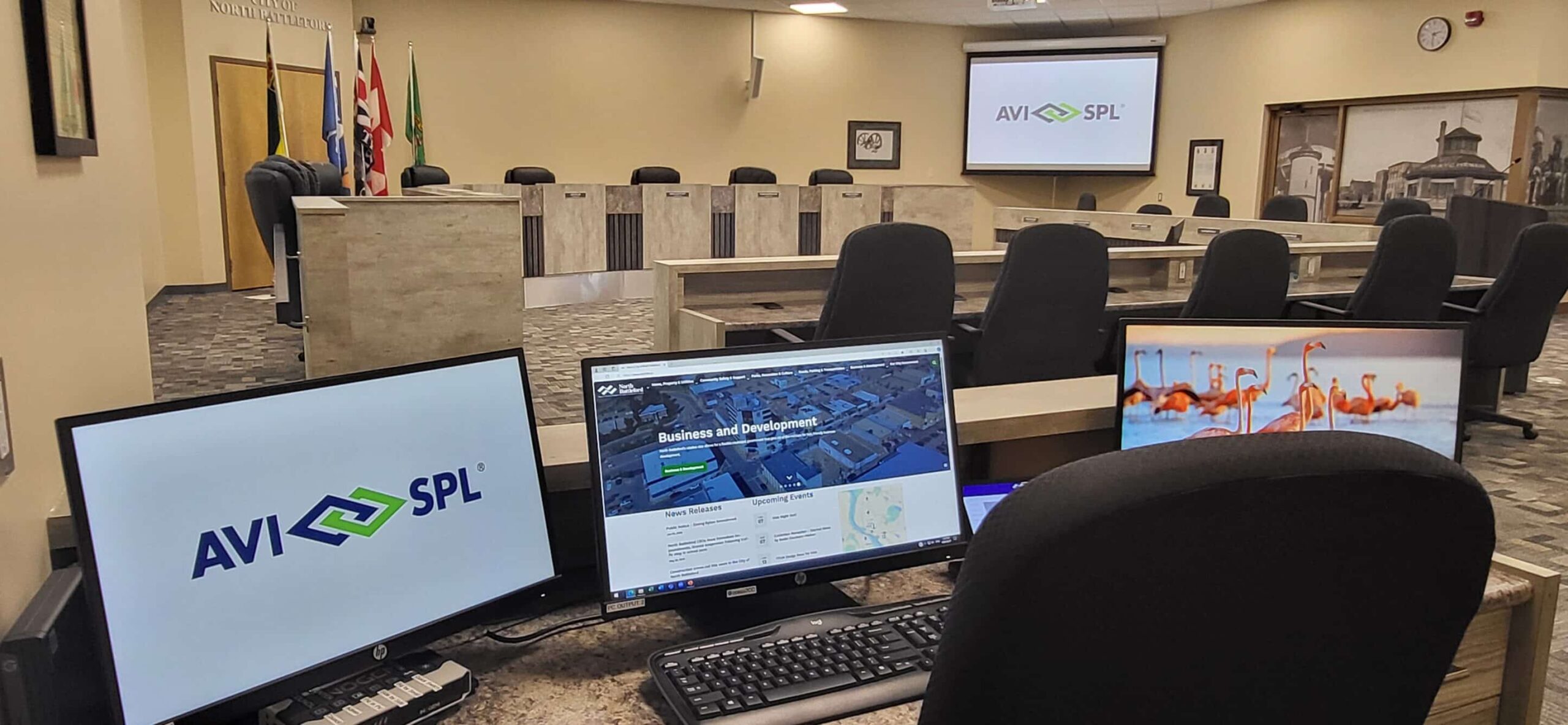 A conference room with desks, chairs, monitors (AVI SPL logo, website, flamingos), flags, and a large screen in the background.