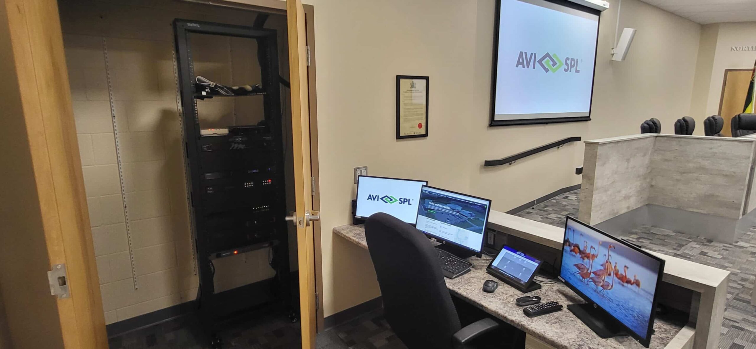 A conference room with AVI-SPL on a screen, desk monitors, control panels, equipment rack in open door, gray chairs around.