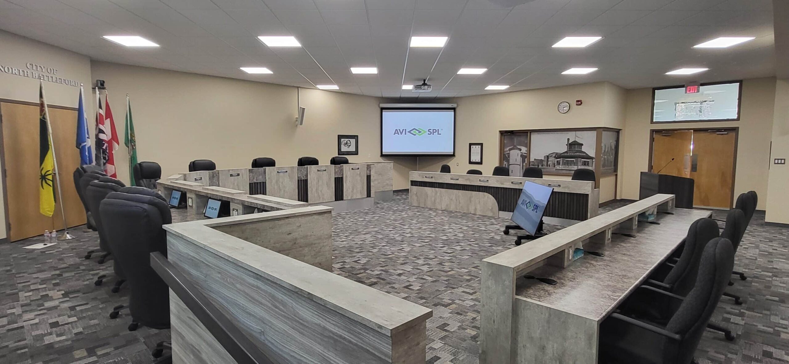 Modern conference room: U-shaped wooden desks, black chairs, screens with logo, flags in corner, mural on back wall, gray pattern carpet.
