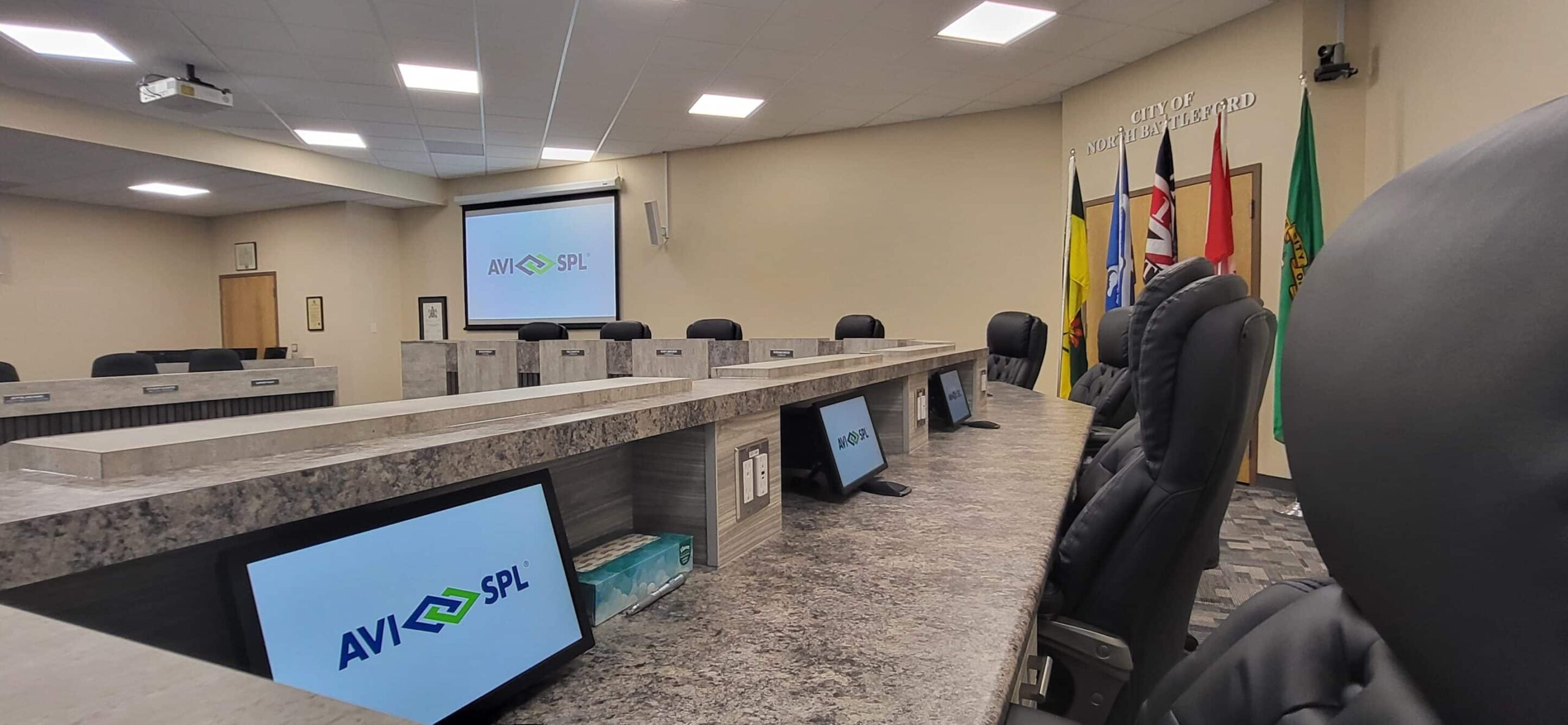 A modern conference room with a large table, cushioned chairs, screens, and flags displaying AVI SPL.