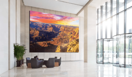 Lobby: large screen with canyon sunset; 3 gray chairs, table on shiny floor; glass doors/windows for light; plant nearby.