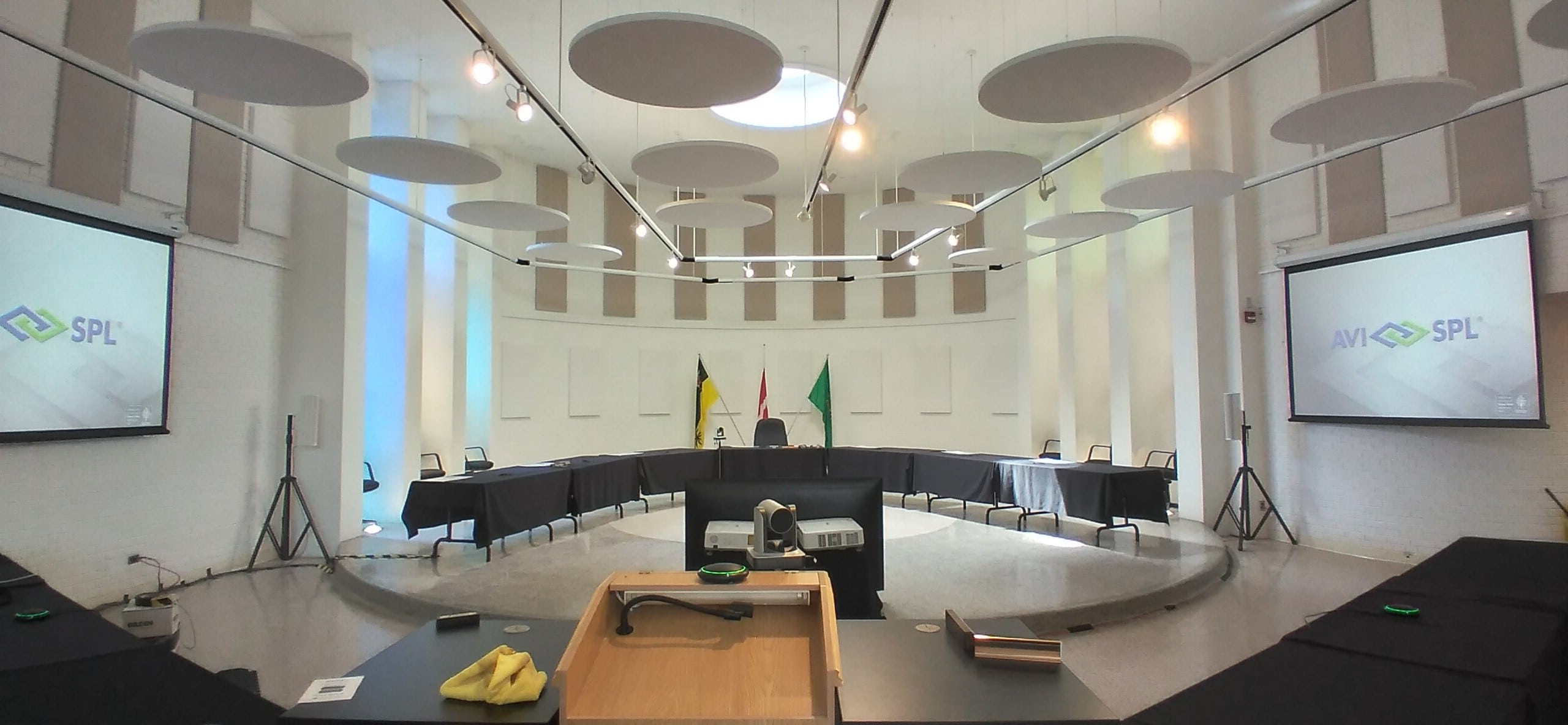 Modern conference room with high ceilings, U-shaped tables, AVI-SPL on screens, flags at front. Circular acoustic panels hang above.