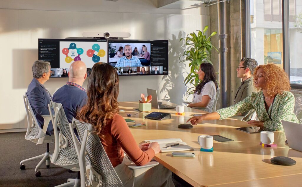 A group sits around a conference table, engaged in a video call. Screen shows participants; room is bright with plants and laptops.