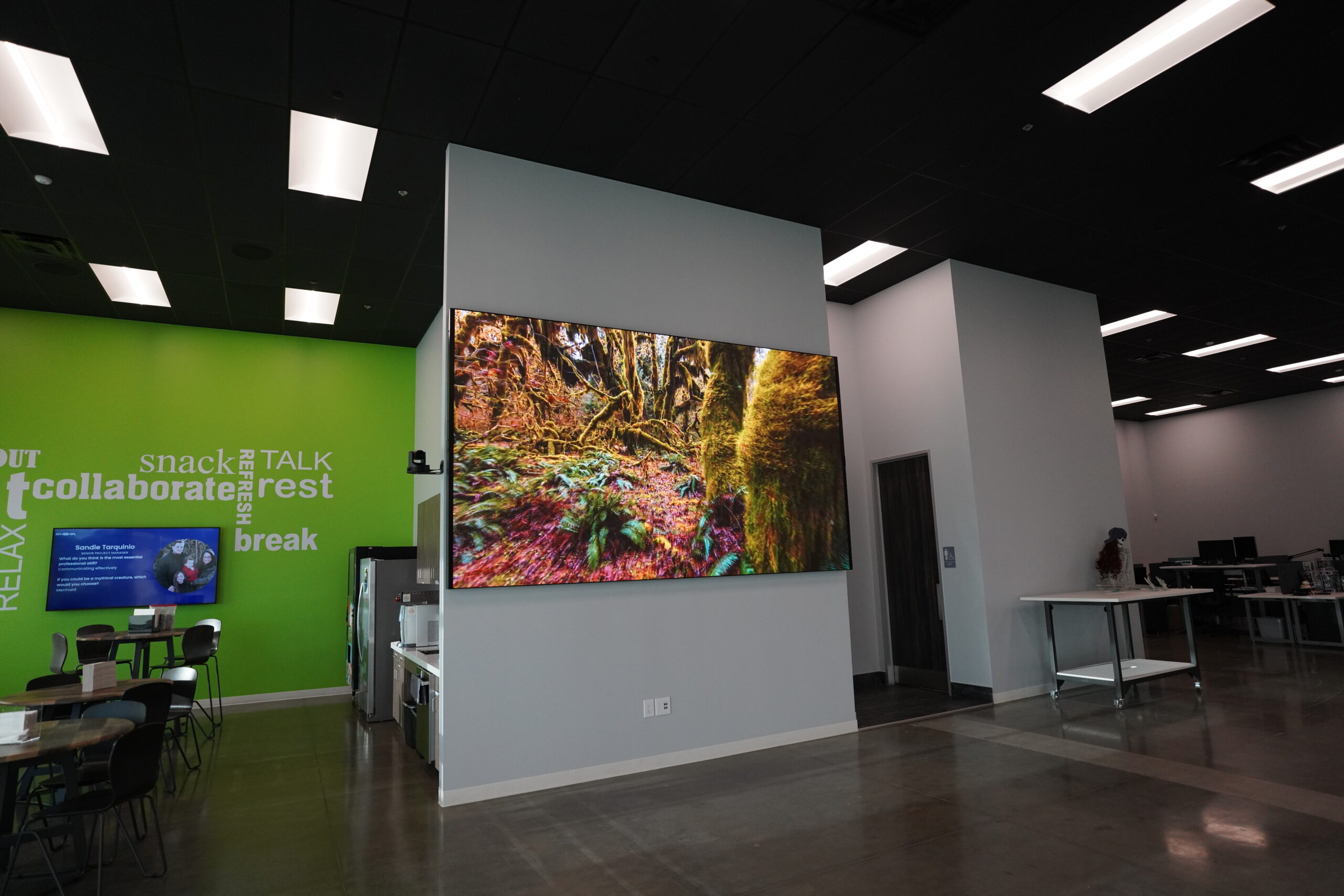Modern office with large screen showing nature on white wall; green wall reads collaborate, snack, break. Tables, chairs, person.