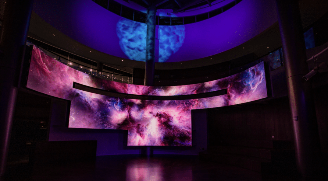 A curved screen shows a vibrant galaxy in swirling colors; a purple ceiling light casts an ambient glow, filling the dark room.